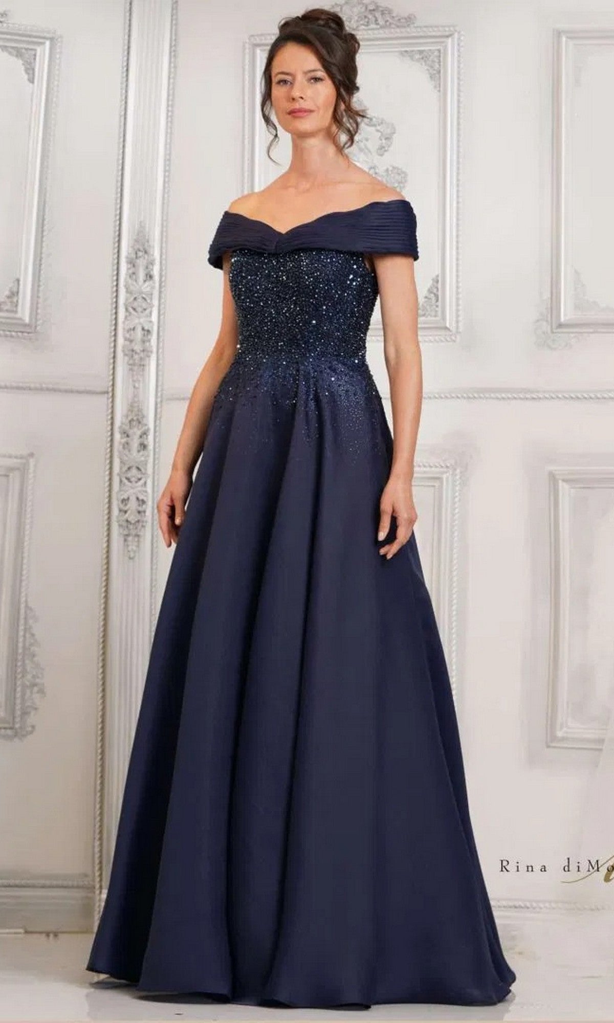 A woman poses elegantly in front of an ornate white wall, wearing the Rina Di Montella RD3102 long formal dress in navy with an off-the-shoulder beaded bodice and a floor-length A-line skirt.
